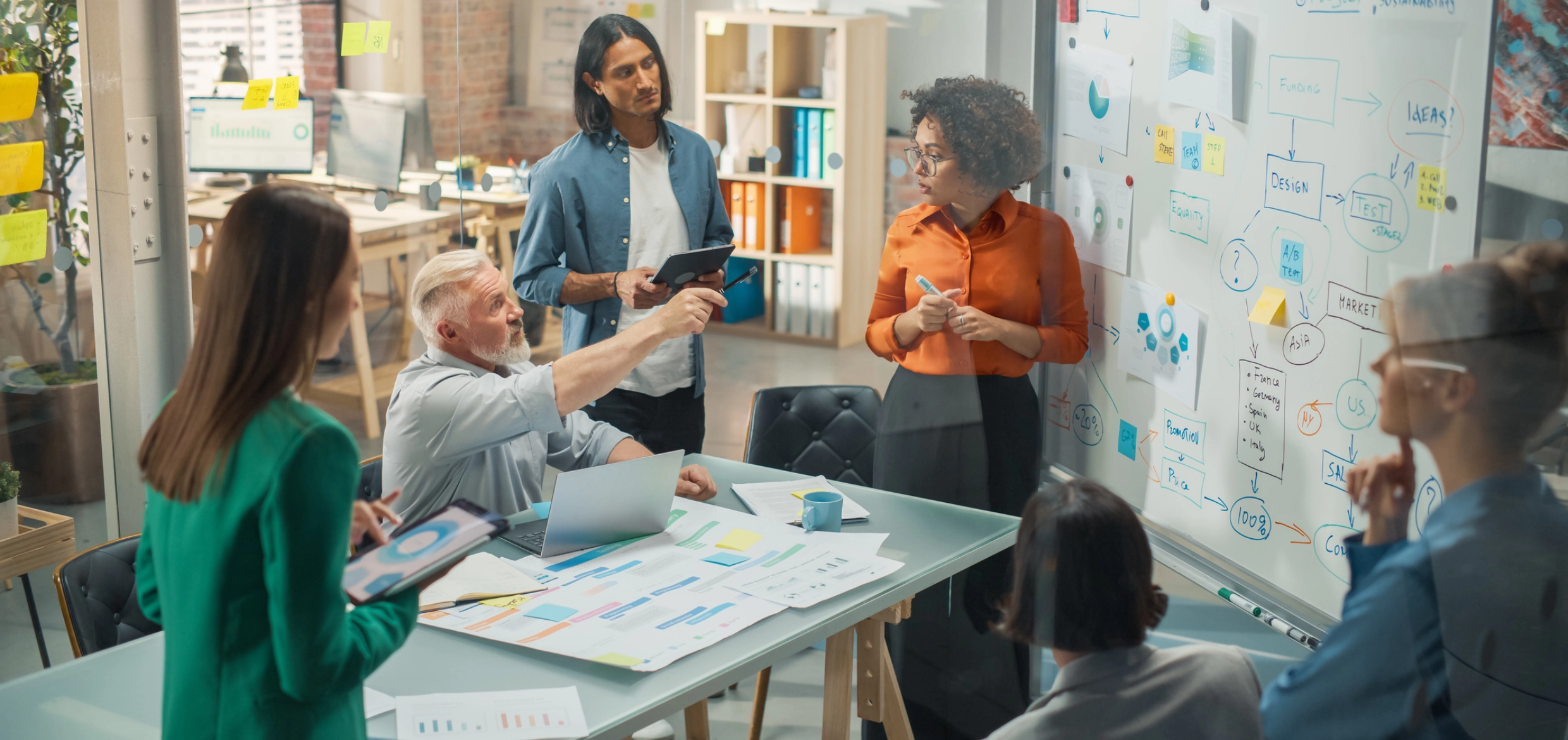 A group of people in a meeting room, discussing and pointing at a whiteboard filled with diagrams and notes. Papers and a laptop are on the table.