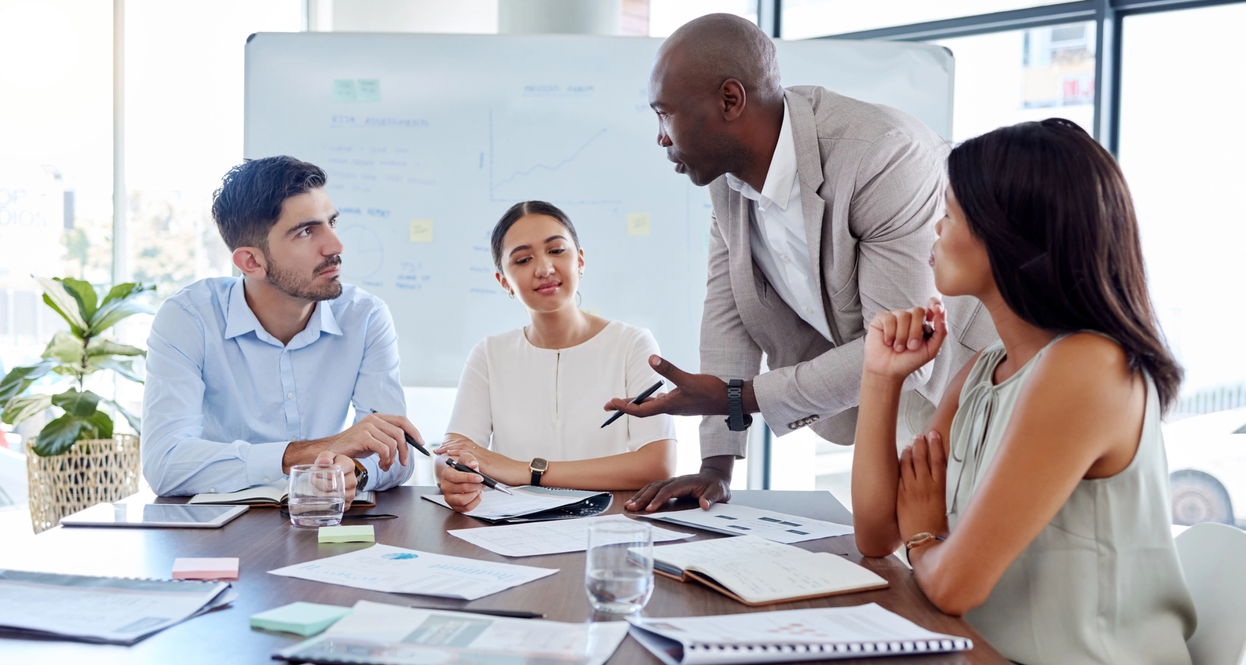 Four people in a meeting room having a discussion around a table with documents, a laptop, and a whiteboard in the background.