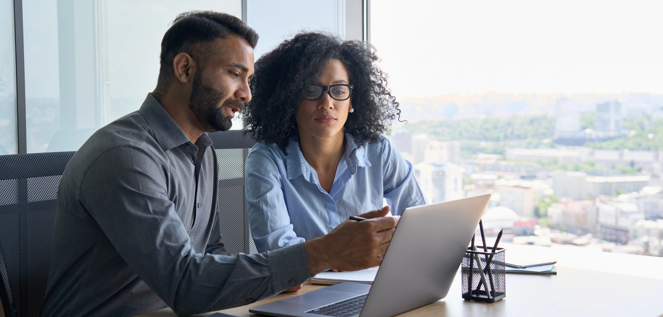 Two people in a modern office look at a laptop screen together. One is pointing at the screen while the other listens attentively. A view of a cityscape is visible through the large window.